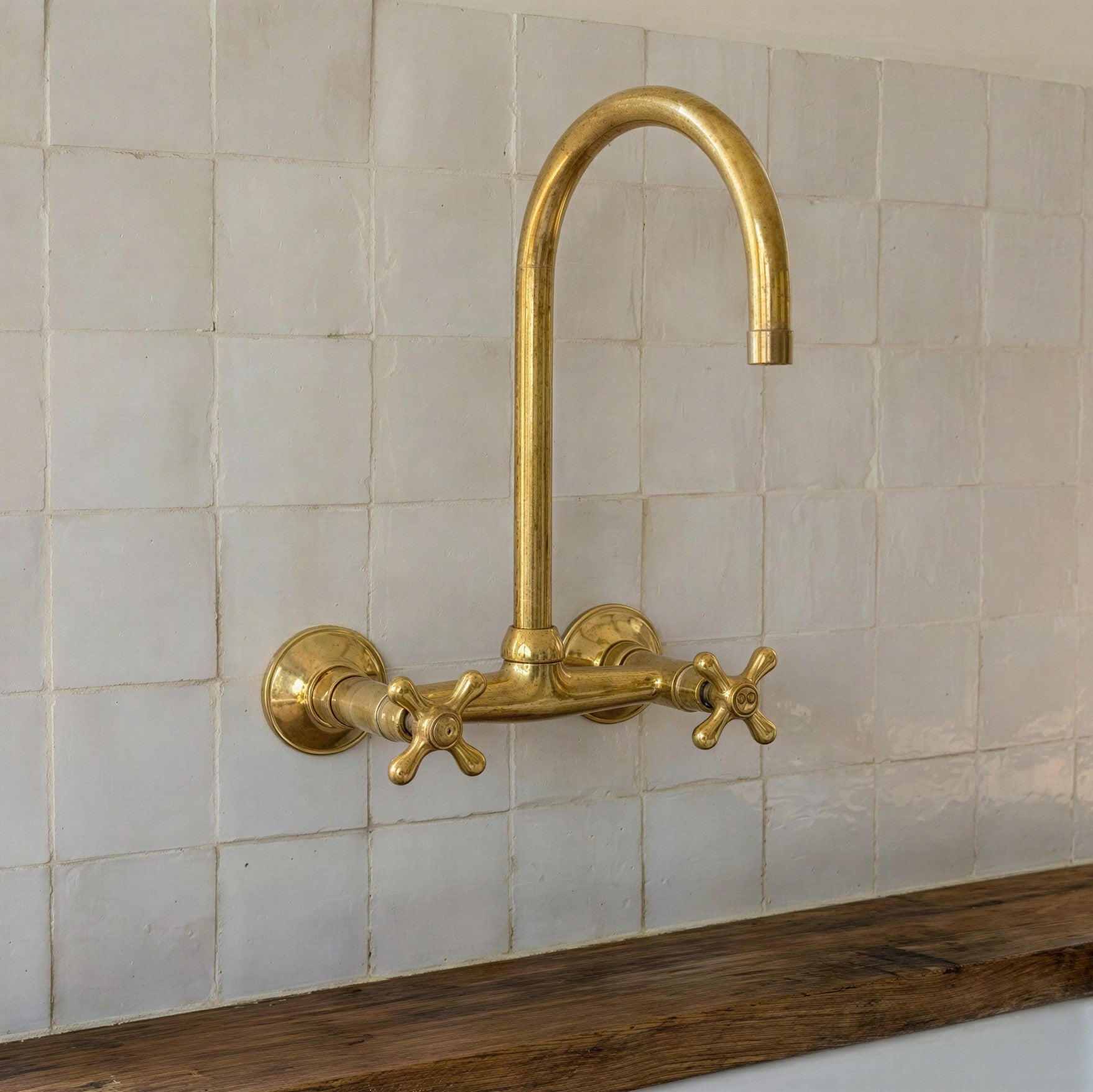 A polished brass wall-mounted faucet with a high-arc spout and two cross handles, installed against a white tiled wall above a dark wooden ledge.