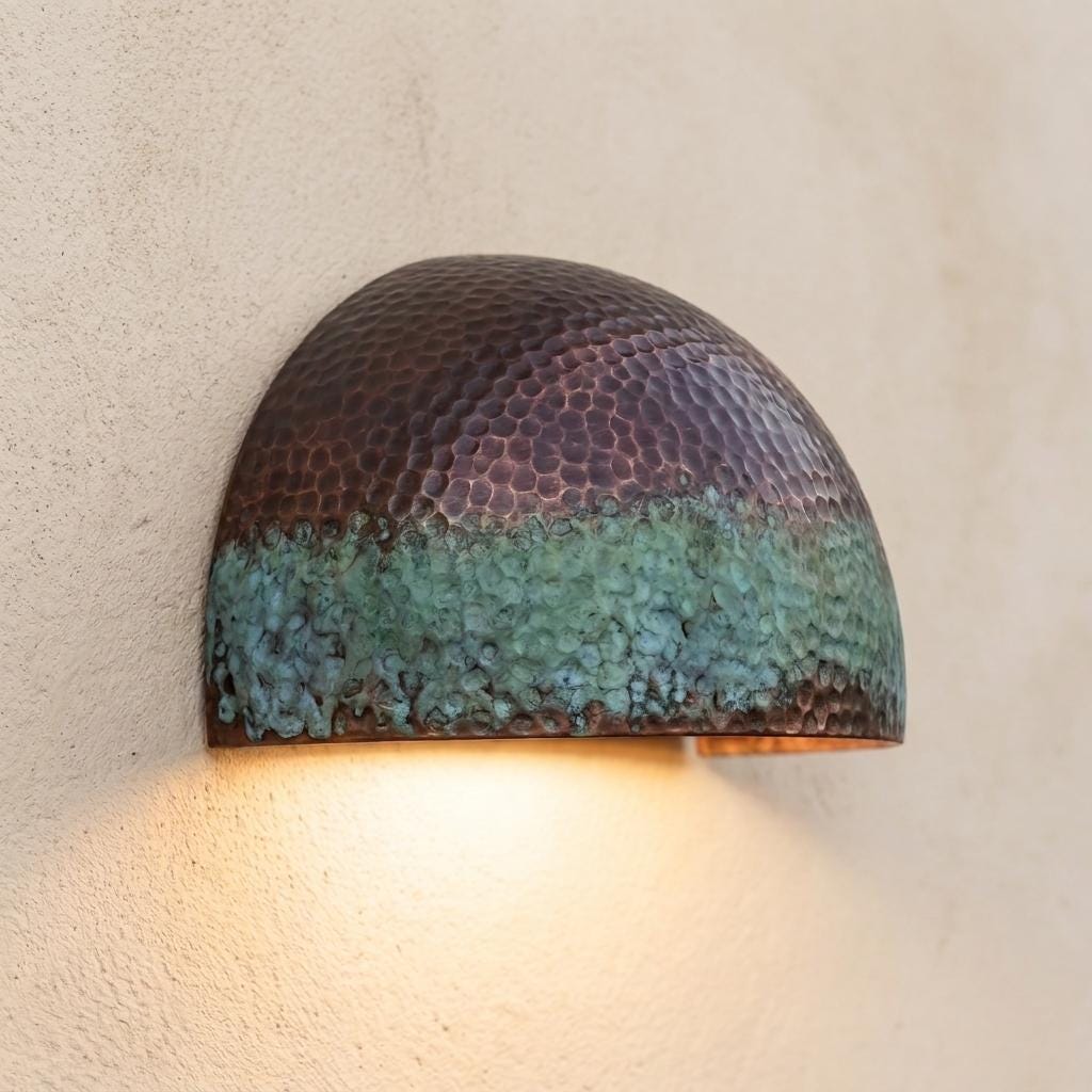 A dome-shaped wall sconce with a hammered metallic finish, featuring a bronze upper section and a textured turquoise-green lower section, casting a warm light against a textured beige wall.