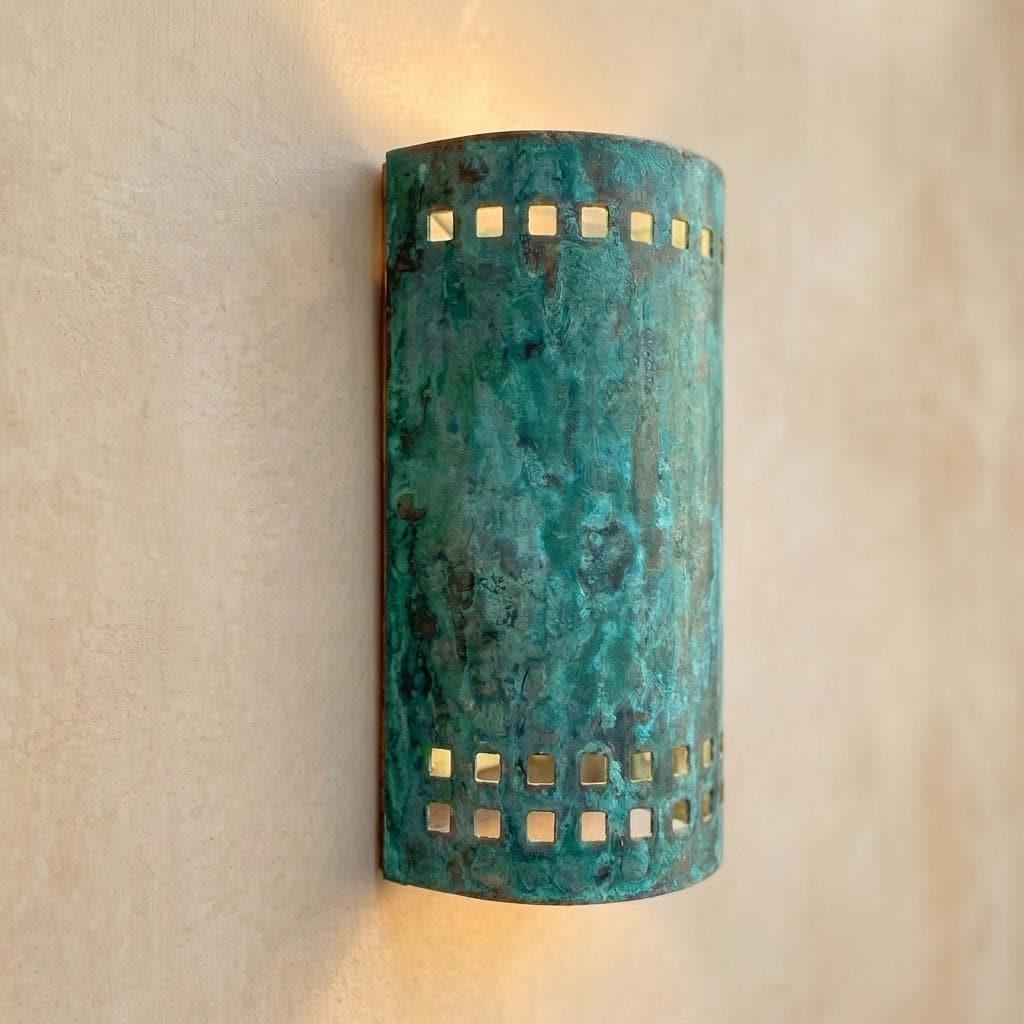 A cylindrical wall sconce with a distressed turquoise and green patina finish, featuring square cutout details on the sides, mounted on a textured beige wall and casting a warm light.
