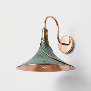 A wall-mounted sconce with a copper-toned metal arm and a conical shade featuring a distressed green patina finish and a polished copper interior.