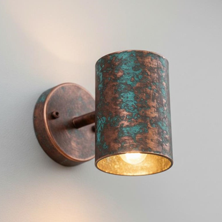 A cylindrical wall sconce with a distressed copper and teal-green patina finish, featuring a warm gold interior and a lit bulb, mounted on a plain light gray wall.