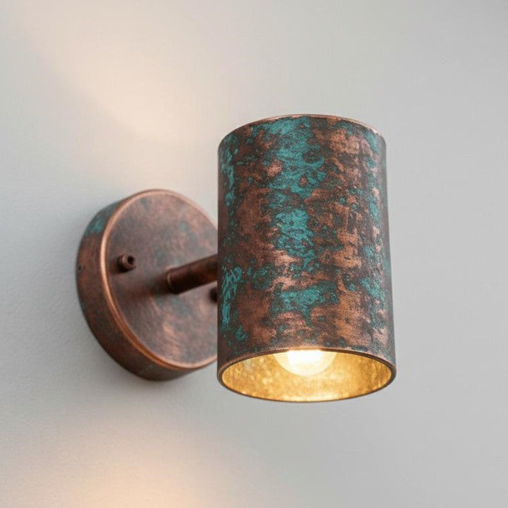 A cylindrical wall sconce with a distressed copper and teal-green patina finish, featuring a warm gold interior and a lit bulb, mounted on a plain light gray wall.