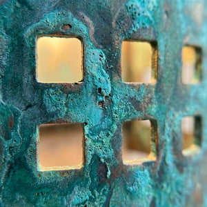 Close-up of a textured metal surface with a turquoise and green patina finish, featuring a grid pattern of square cutouts showing a warm blurred background.