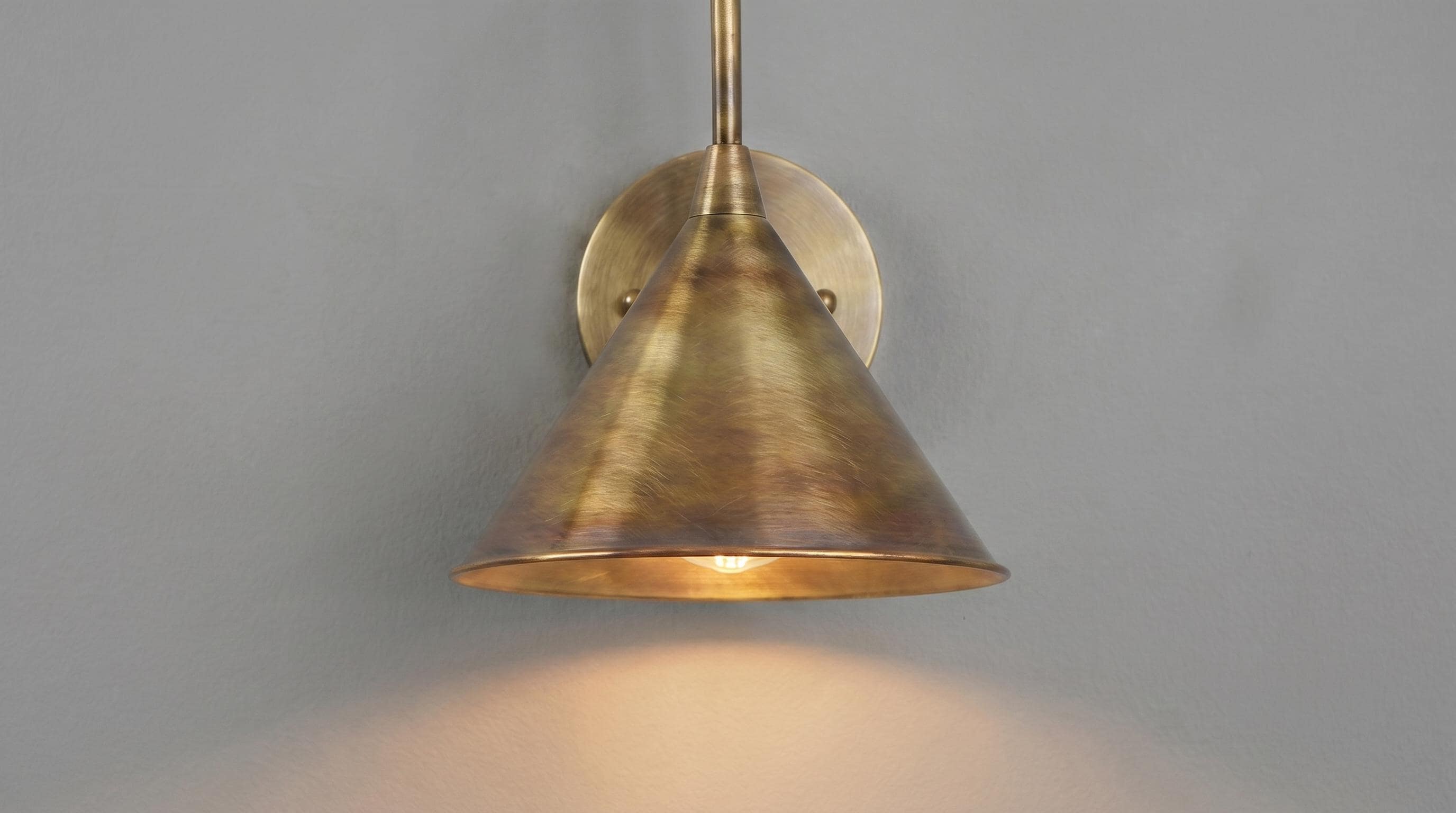 A brass wall sconce with a conical shade and circular wall mount, shown illuminated against a plain gray wall.