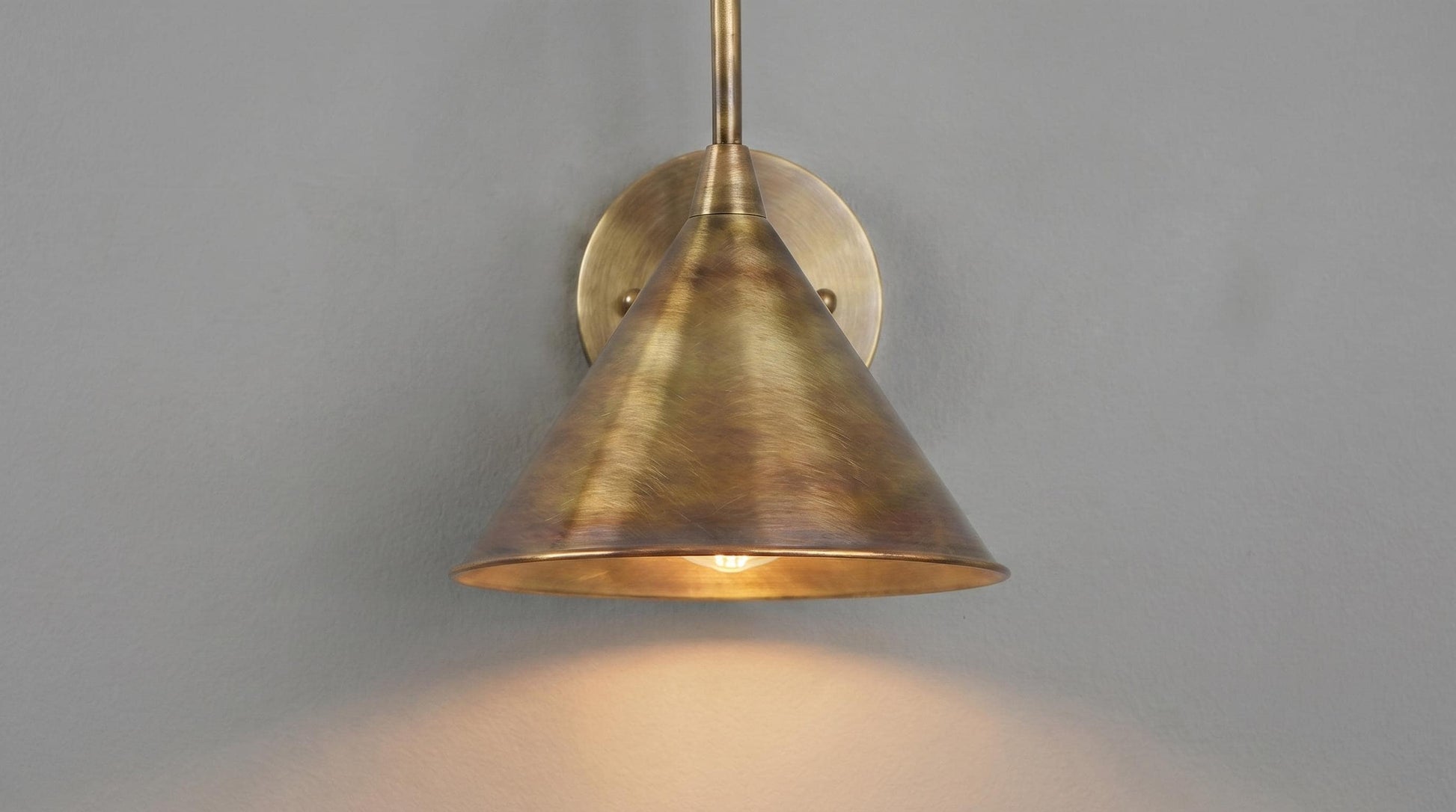 A brass wall sconce with a conical shade and circular wall mount, shown illuminated against a plain gray wall.