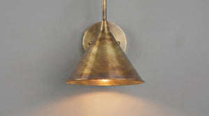 A brass wall sconce with a conical shade and circular wall mount, shown illuminated against a plain gray wall.