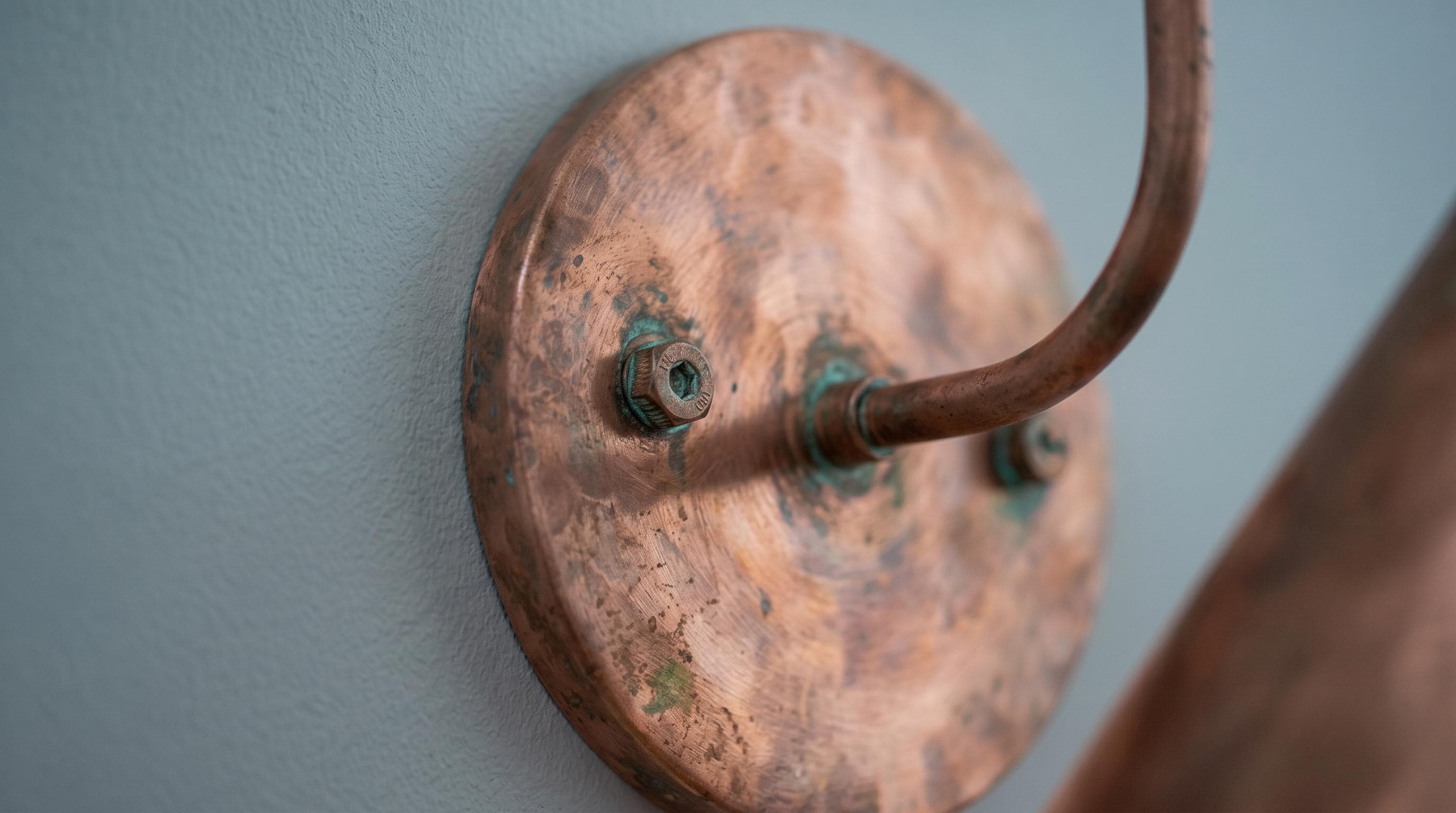 A close-up of a rustic copper wall sconce with a round disc base and a curved hook arm, mounted on a textured light blue-gray wall.