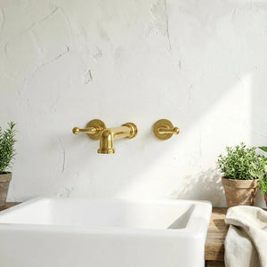 A wall-mounted faucet set in a warm brushed gold finish installed above a white sink, with a textured white wall, potted plants, and a folded towel in the background.
