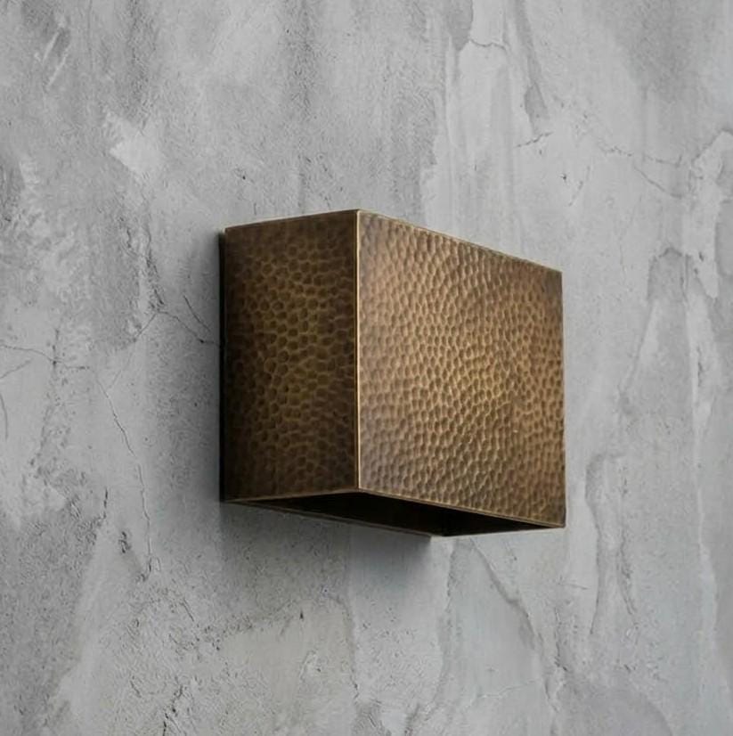 A square wall-mounted light fixture with a hammered metallic bronze finish, mounted on a textured gray concrete wall.