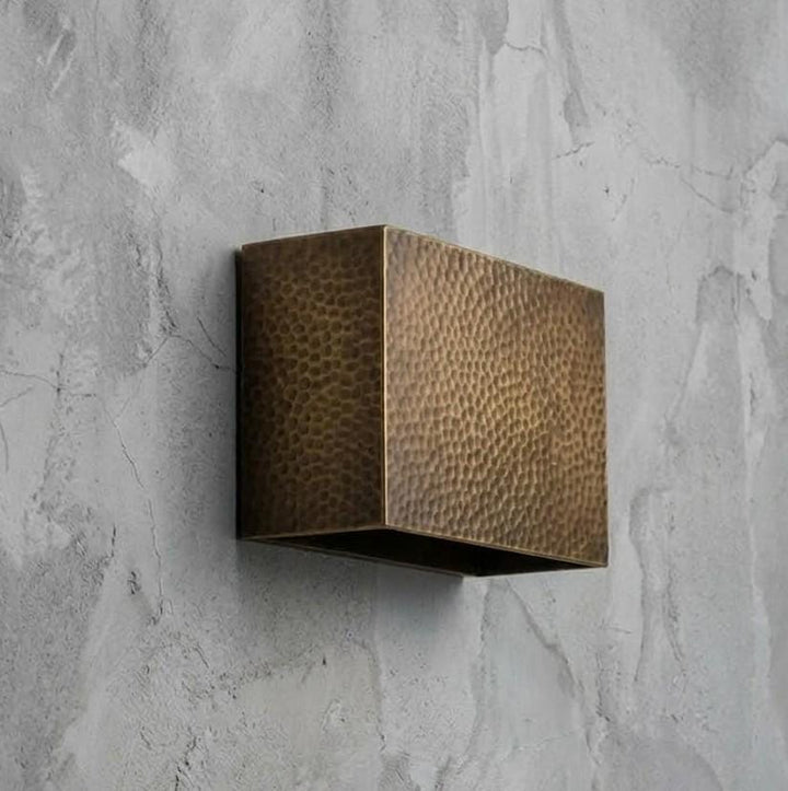 A square wall-mounted light fixture with a hammered metallic bronze finish, mounted on a textured gray concrete wall.