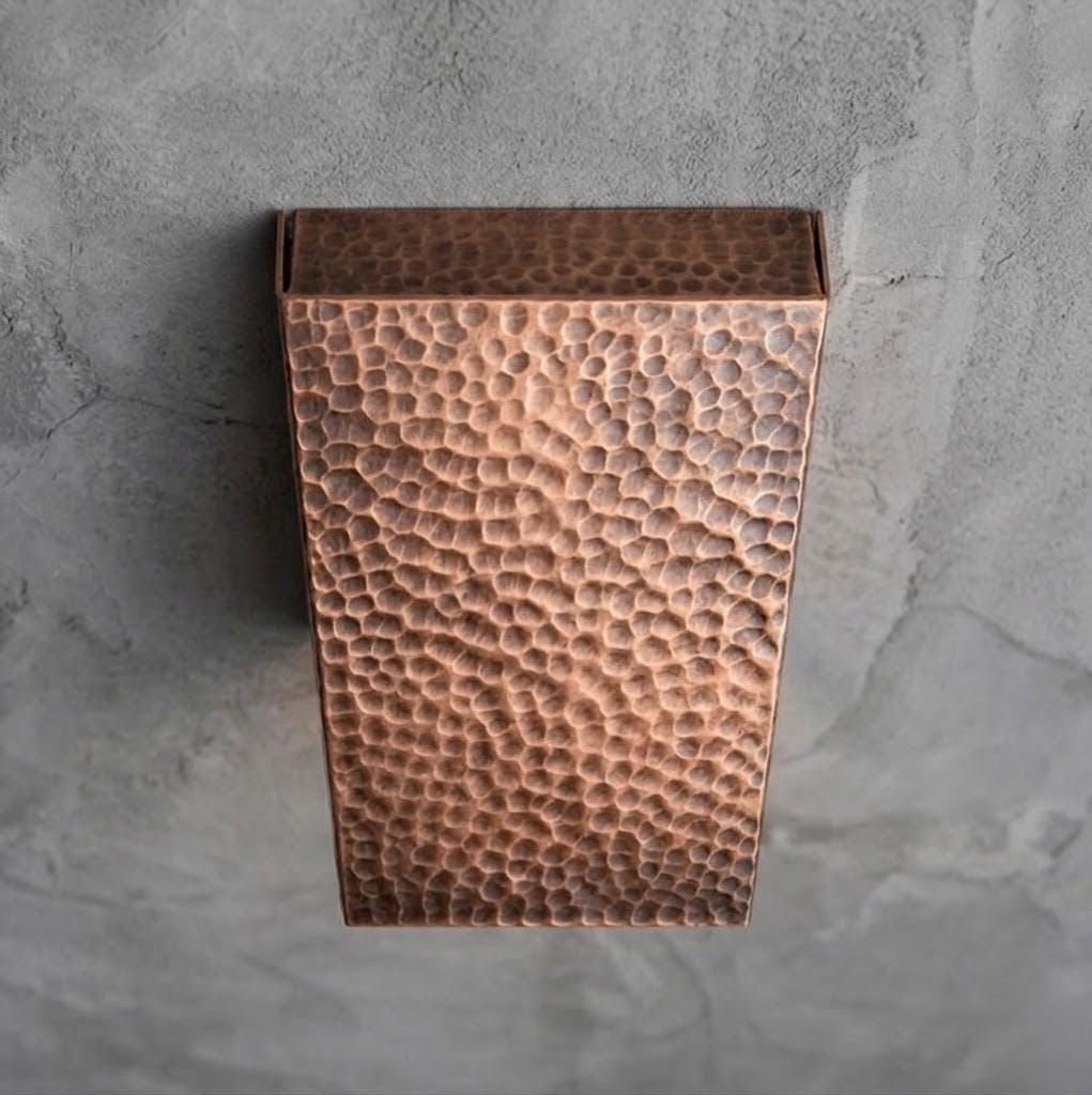 A rectangular hammered copper wall sconce mounted on a textured gray concrete wall.