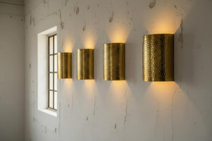 Four cylindrical hammered metal wall sconces in a warm gold finish, mounted in a row on a textured white wall next to a window.