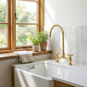 A modern kitchen sink setup with a tall arched brass faucet, white countertop, white subway tile backsplash, and a wooden window frame with potted green plants on the sill.