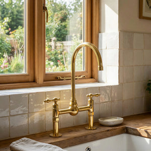 Handmade unlacquered brass bridge kitchen faucet with high-arc gooseneck spout and classic lever handles mounted on a rustic wood countertop over a fireclay sink.