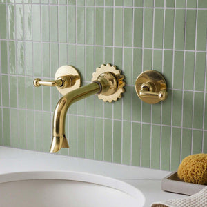 Gold faucet and handles on a green tiled wall with a sink and sponge in the foreground.
