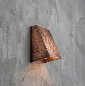 A modern geometric wall sconce with a hammered copper finish, mounted on a textured gray concrete wall and casting a warm light downward.
