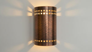 A cylindrical wall sconce with a textured bronze finish and square cutouts, casting warm light and shadow patterns on a light gray wall.