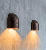 Two modern dome-shaped wall sconces with a hammered dark metal finish, mounted on a textured gray concrete wall and casting a warm yellow light downward. A wooden console table is visible in the foreground with a small vase and book.