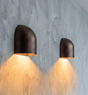 Two modern dome-shaped wall sconces with a hammered dark metal finish, mounted on a textured gray concrete wall and casting a warm yellow light downward. A wooden console table is visible in the foreground with a small vase and book.