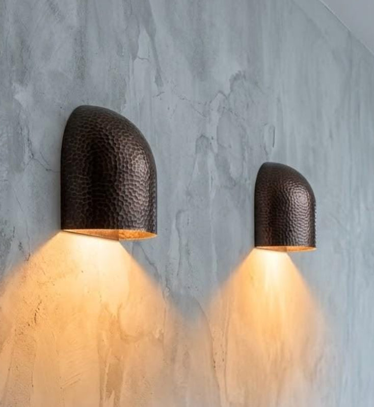 Two modern dome-shaped wall sconces with a hammered dark metal finish, mounted on a textured gray concrete wall and casting a warm yellow light downward. A wooden console table is visible in the foreground with a small vase and book.