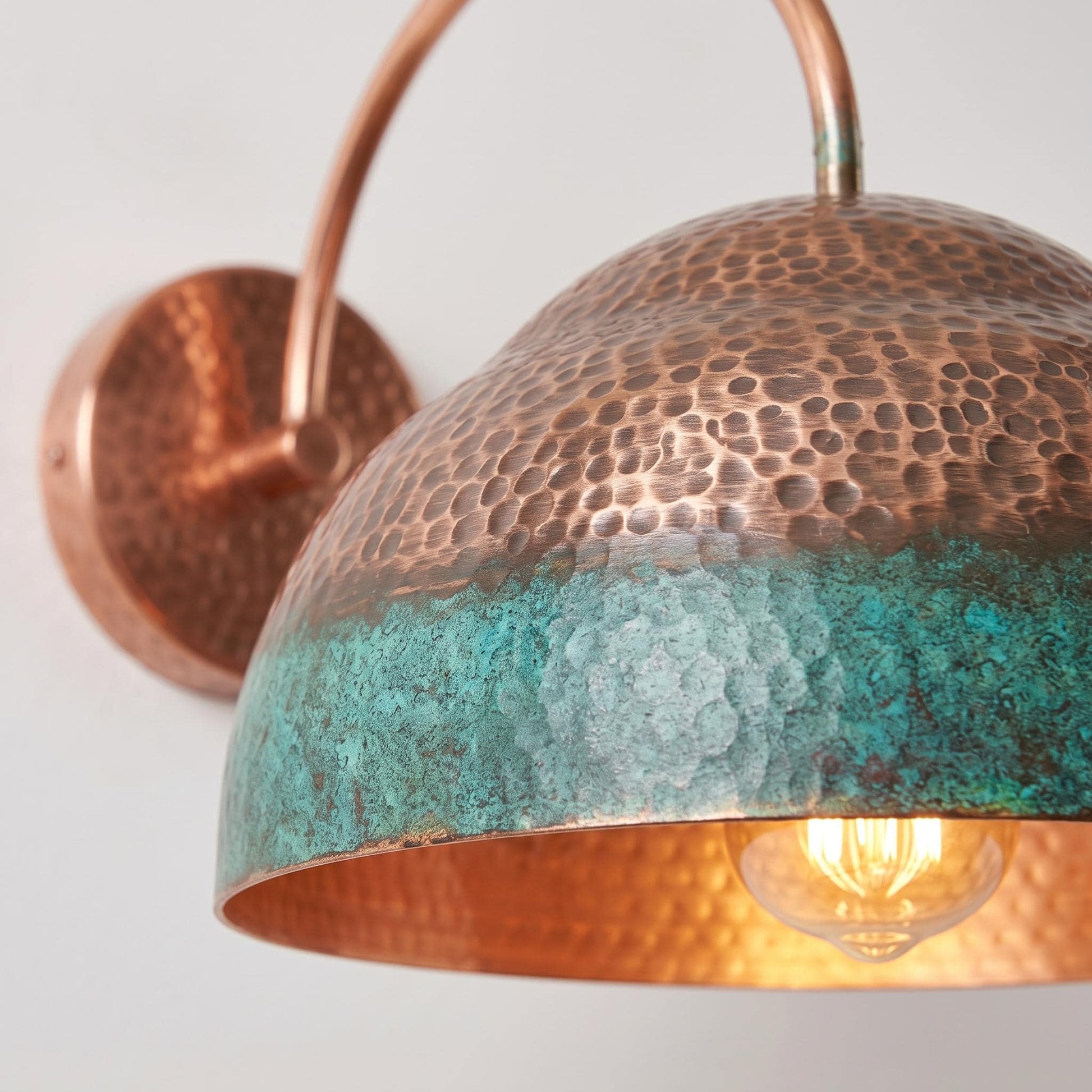 Close-up of a hammered copper dome wall sconce with a turquoise oxidized finish and a glowing light bulb inside, mounted on a plain light background.
