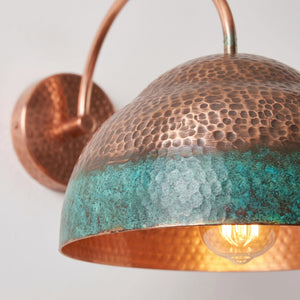 Close-up of a hammered copper dome wall sconce with a turquoise oxidized finish and a glowing light bulb inside, mounted on a plain light background.