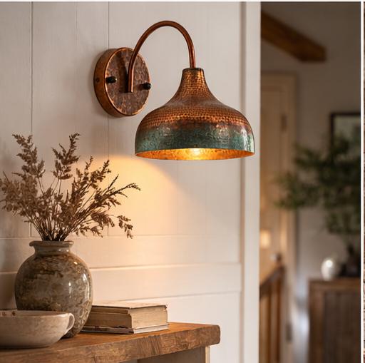 A rustic copper and teal hammered metal wall sconce mounted on a white paneled wall above a wooden console table with a vase and books, casting a warm glow.