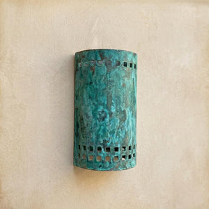 A cylindrical wall sconce with a distressed turquoise and green patina finish, featuring square cutout details near the top and bottom, mounted on a textured beige wall.