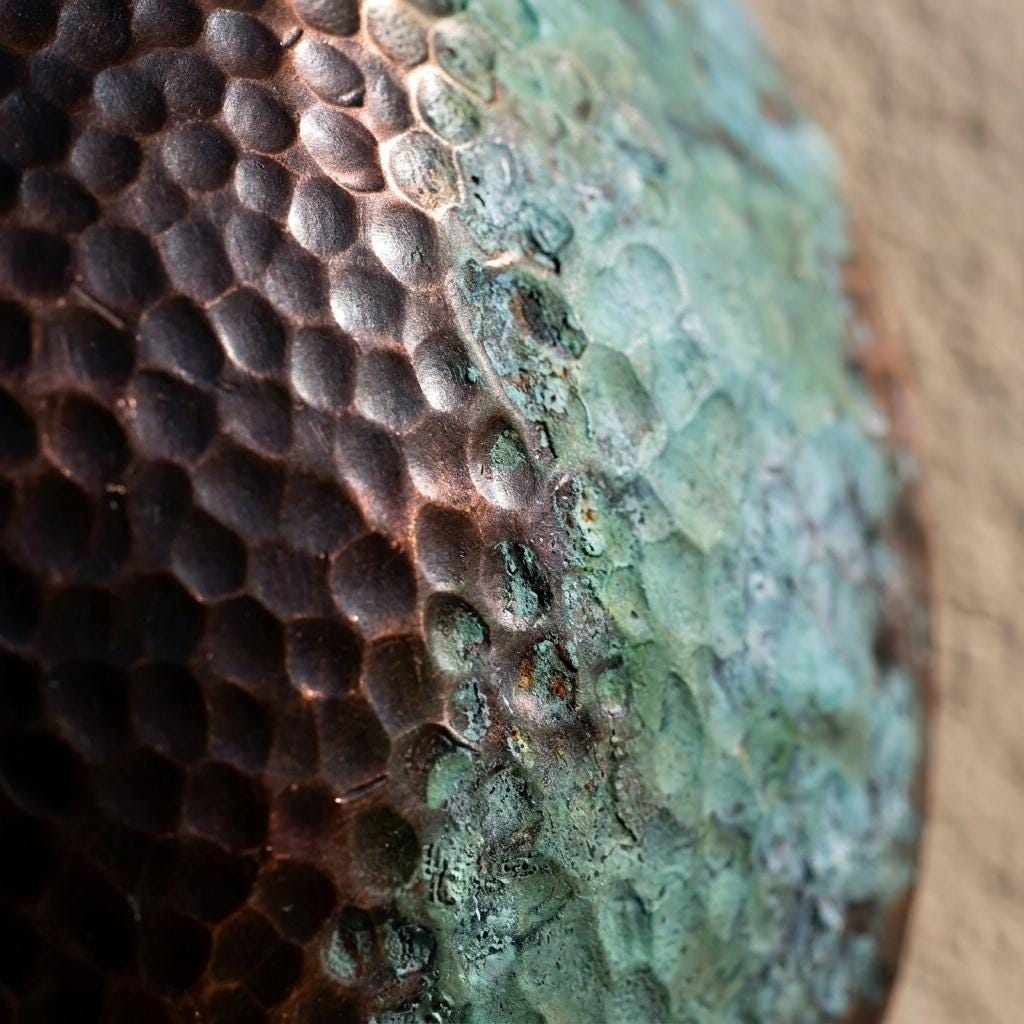 Macro close-up of a textured metallic surface with a hammered hexagonal pattern, transitioning from dark bronze-brown on the left to a greenish patina on the right, against a beige background.