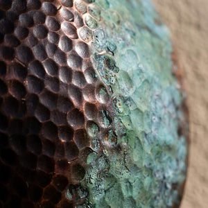 Macro close-up of a textured metallic surface with a hammered hexagonal pattern, transitioning from dark bronze-brown on the left to a greenish patina on the right, against a beige background.