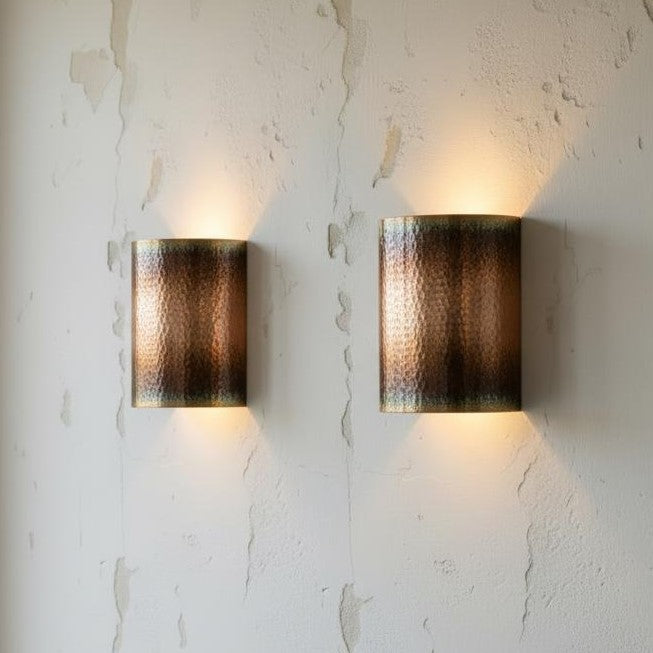 Two cylindrical wall sconces with a hammered metallic finish, mounted on a textured off-white plaster wall and casting a warm light downward.