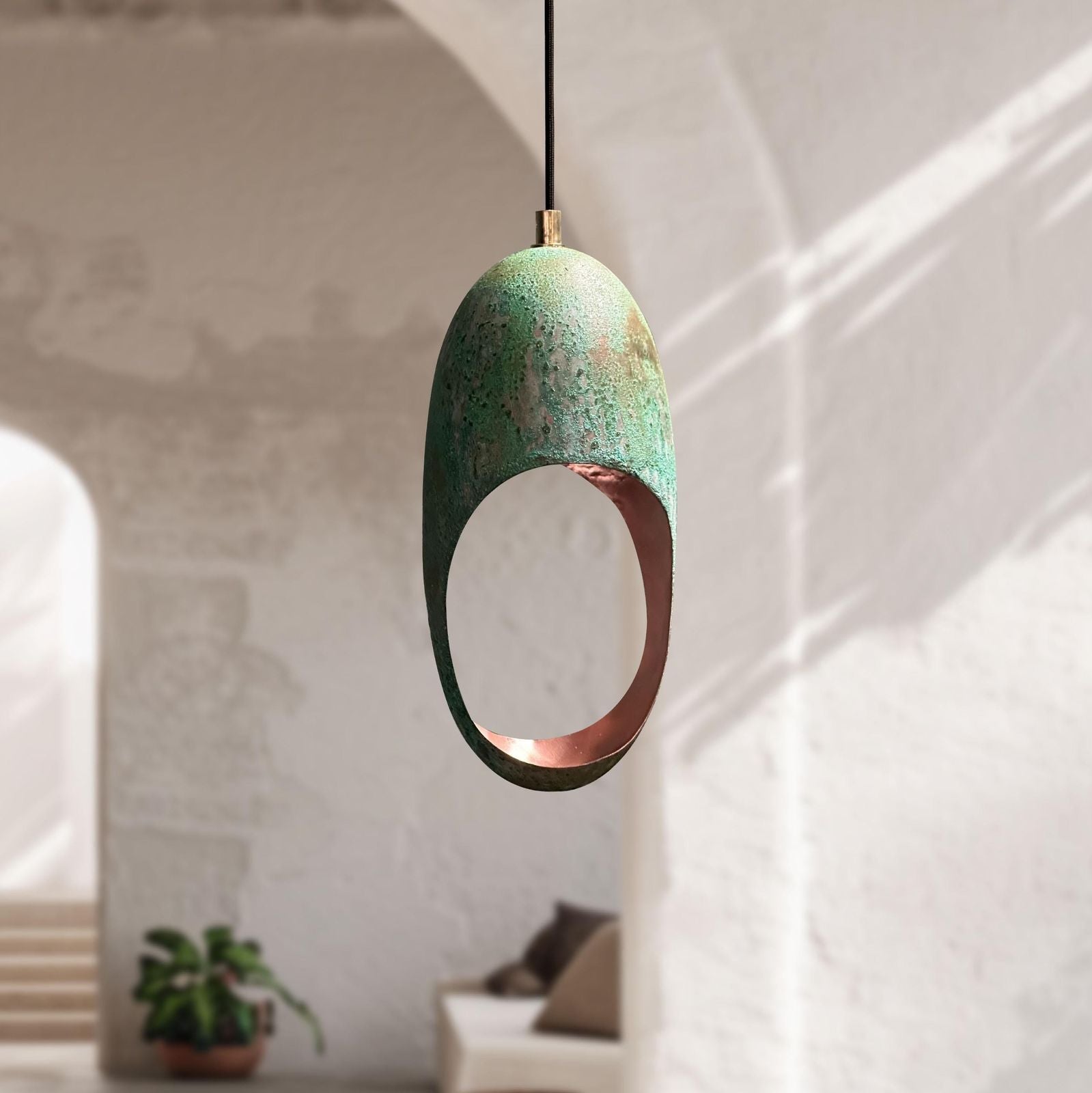 An organic-shaped pendant light with a distressed green patina finish and a copper-toned interior, hanging in a softly lit modern interior with arches and a blurred sofa in the background.