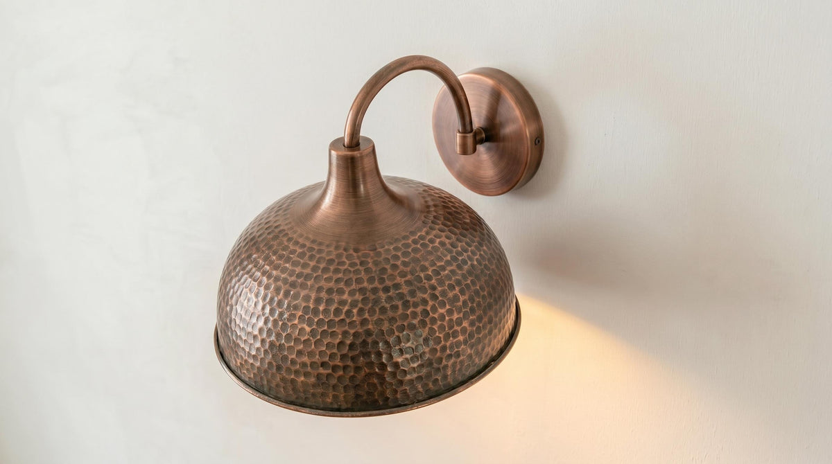 A hammered copper dome wall sconce with a curved arm and round wall mount, shown illuminated against a plain off-white wall.