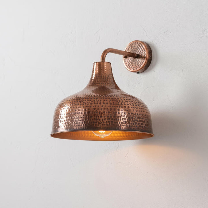 A hammered copper dome wall sconce with a warm glowing light bulb, mounted on a textured off-white wall.