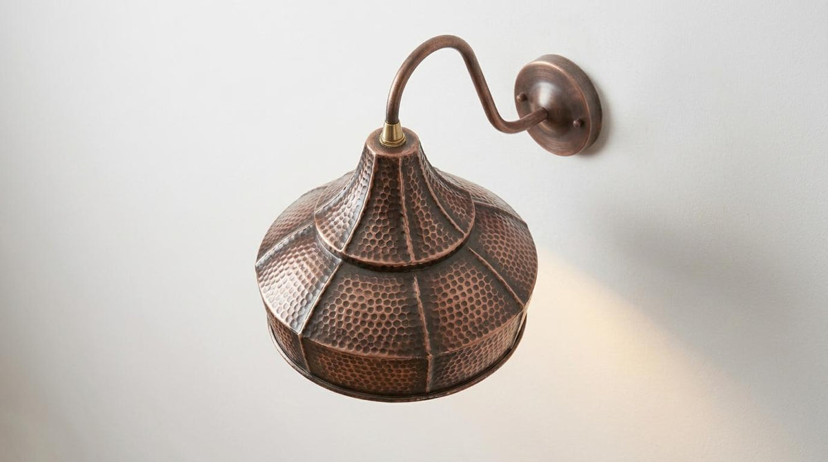 A copper-toned metal wall sconce with a textured, dome-shaped shade and a curved arm, mounted on a plain light gray wall with a warm light shining downward.