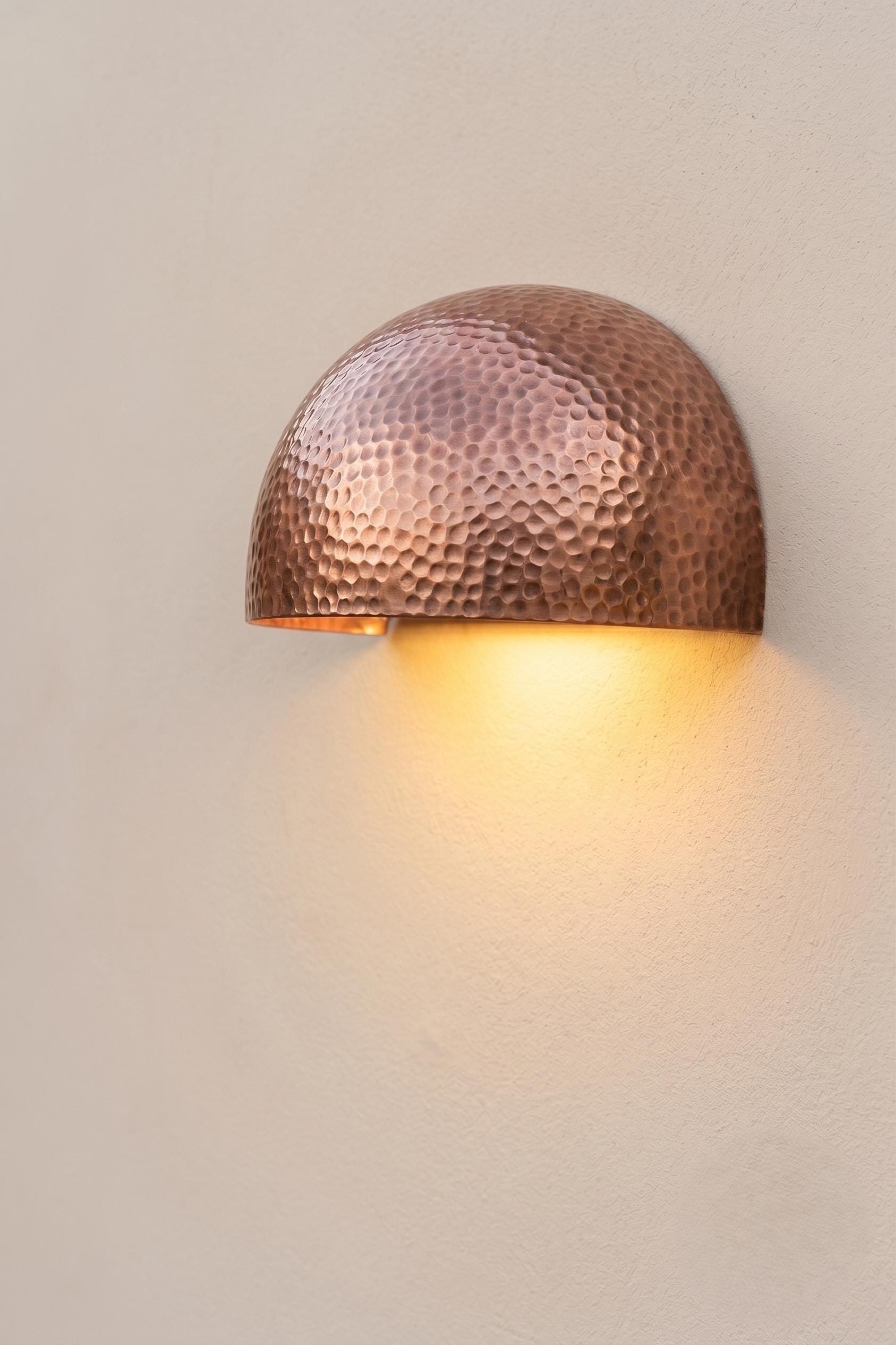 A hammered copper dome wall sconce casting a warm yellow light against a textured beige wall.