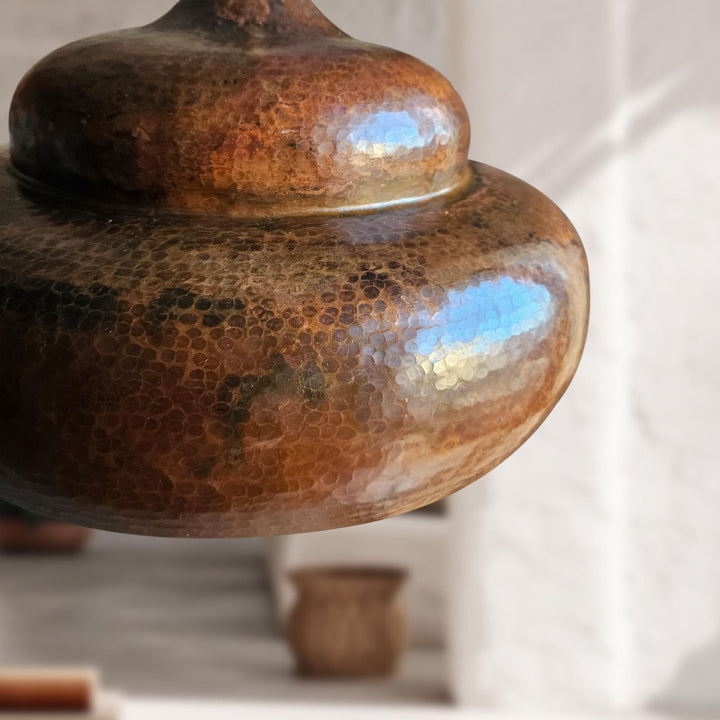 A close-up of a hammered metal pendant light shade with a warm coppery-brown finish and subtle blue iridescent reflections, shown against a softly blurred white interior background.