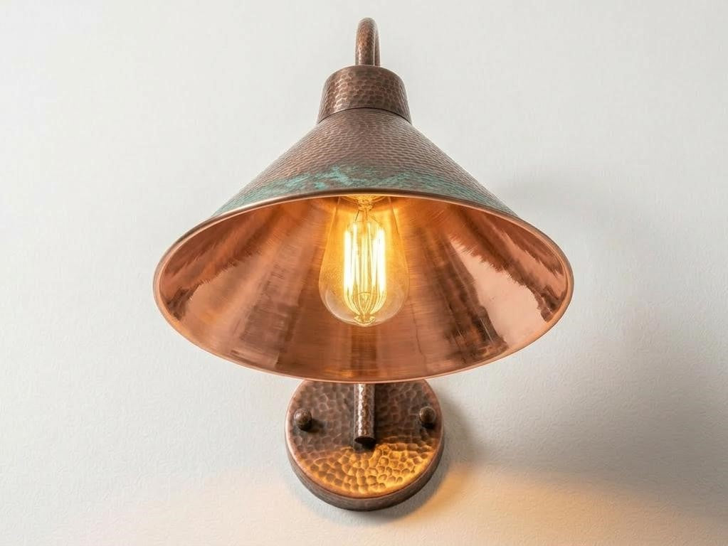 A rustic copper wall sconce with a hammered metal base and a conical shade, glowing with a warm light against a plain off-white background.
