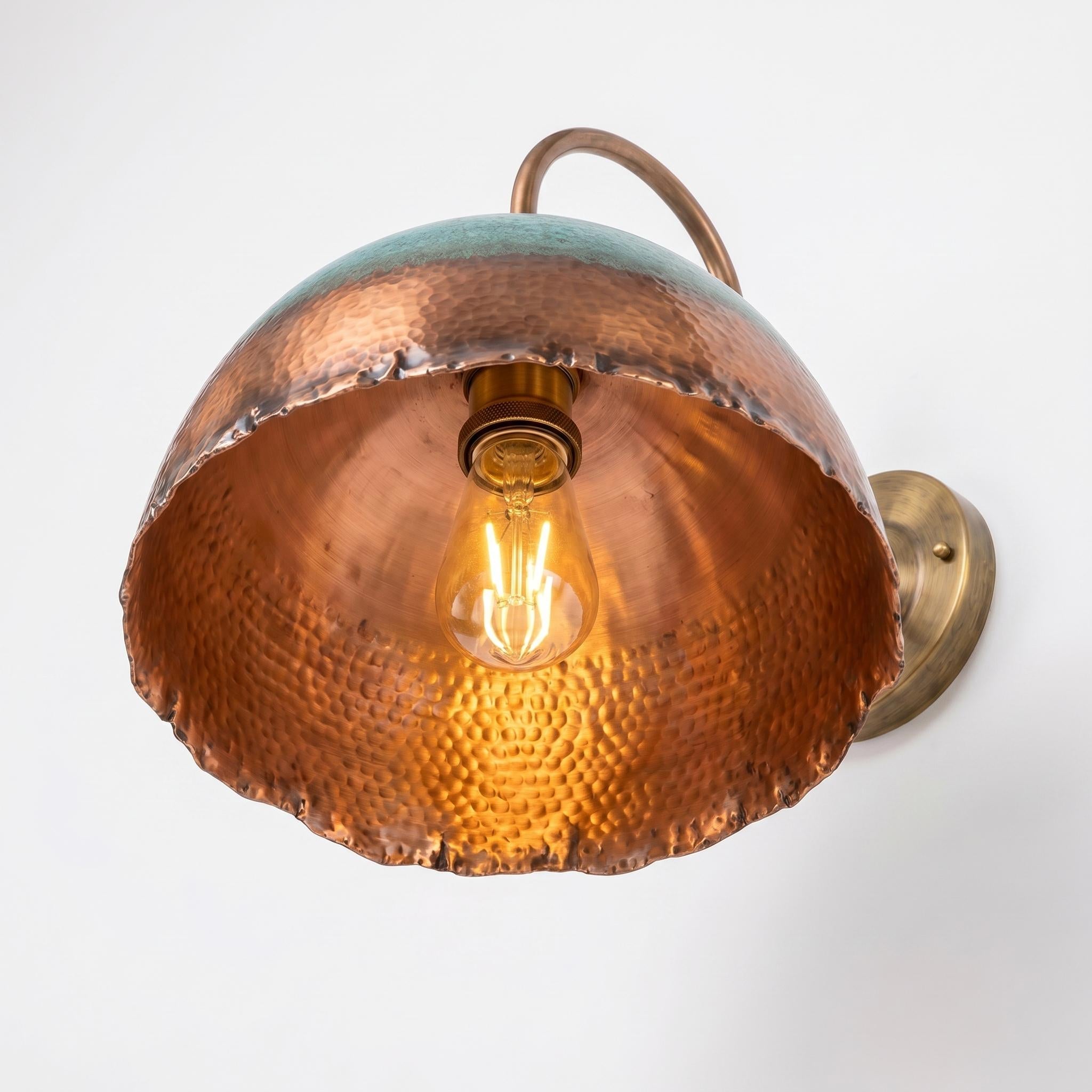 A rustic copper dome wall sconce with a hammered texture, a warm glowing light bulb inside, and a brass wall mount on a plain white background.