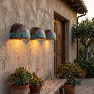 Three dome-shaped outdoor wall sconces with a hammered metallic finish and turquoise-green patina, mounted on a stucco wall above potted succulents and a wooden bench.