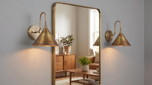 A tall rectangular wall mirror with a gold-toned frame, mounted on a light gray wall between two matching gold sconces. The mirror reflects a warm, cozy living room interior with wood furniture and green plants.