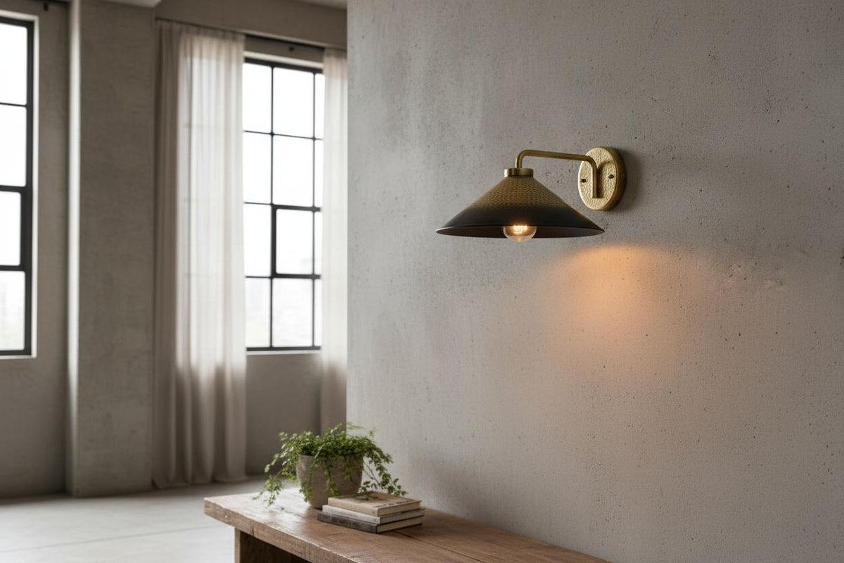 A modern wall sconce with a dark conical shade and a warm brass fixture, mounted on a textured concrete wall in a minimalist room with a wooden bench and windows.
