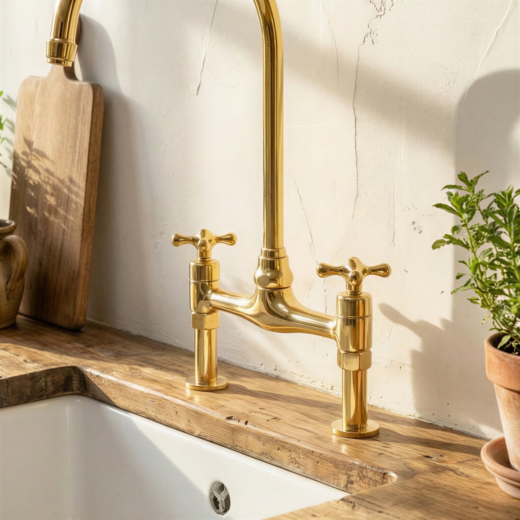 A polished brass bridge faucet with two cross handles installed on a rustic wooden kitchen sink countertop, with a white sink basin, textured cream wall, wooden cutting board, and potted plant nearby.