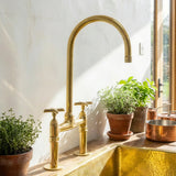 A polished brass kitchen faucet with a high-arc spout and two cross-handle valves, installed on a wooden countertop beside potted plants and a copper pot, with a white plaster wall and window in the background.