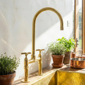 A polished brass kitchen faucet with a high-arc spout and two cross-handle valves, installed on a wooden countertop beside potted plants and a copper pot, with a white plaster wall and window in the background.