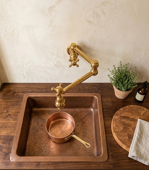 A hammered copper farmhouse-style sink with a matching copper pot, paired with a polished brass wall-mounted pot faucet, set on a dark wood countertop against a textured beige wall.