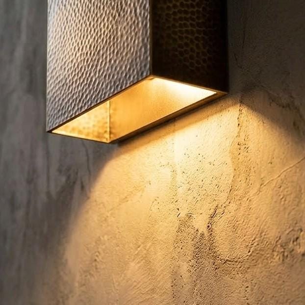 A modern wall-mounted light fixture with a textured hammered metal shade and a warm glowing interior, mounted on a rough concrete wall.