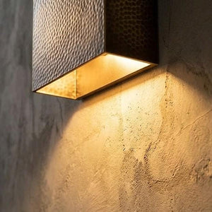 A modern wall-mounted light fixture with a textured hammered metal shade and a warm glowing interior, mounted on a rough concrete wall.