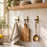 Three solid brass kitchen wall hooks holding a linen towel, cutting board and utensil