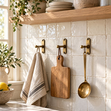 Three solid brass kitchen wall hooks holding a linen towel, cutting board and utensil